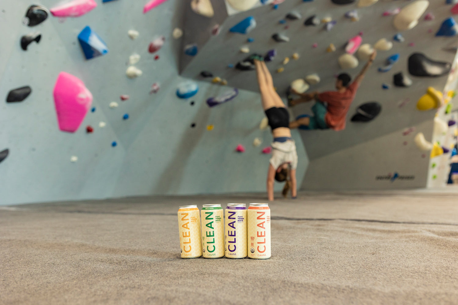 Set in a rocking climbing gym - the background is a rock climbing wall with colorful hand and foot holds. There is a man climbing and a woman doing a handstand in the background. In the foreground is a can of each flavor of CLEAN Cause Non-Carbonated Organic Yerba Mate